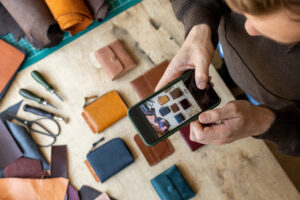 Top view male tanner hands taking photo of exclusive leatherwork accessories on wooden table POV shot. Craftsman photographing handmade wallets use smartphone.